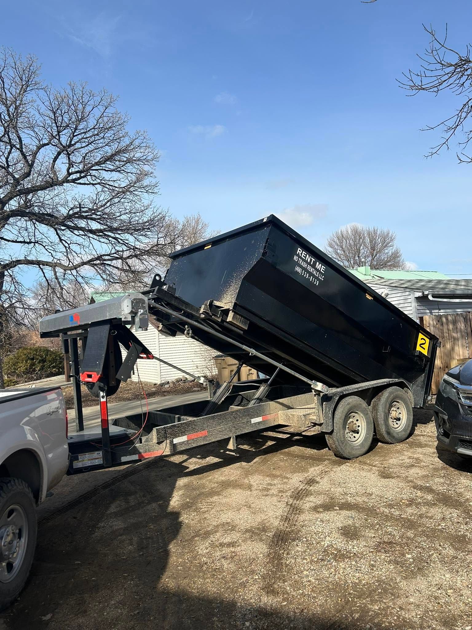 Black dump trailer hitched to a truck in a yard, tilted with its bed raised under a clear blue sky