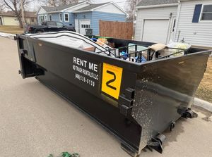 Black rental dumpster on a street beside garages, filled with debris and marked with a yellow number 2.