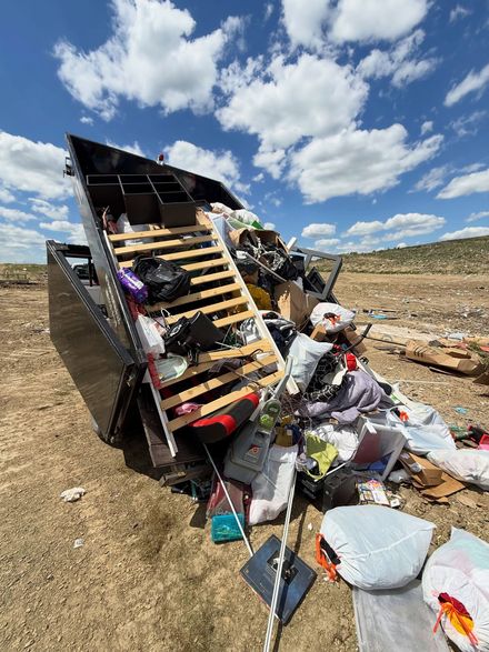 Overturned trash dumpster spilling garbage in a dry field under a blue sky with clouds