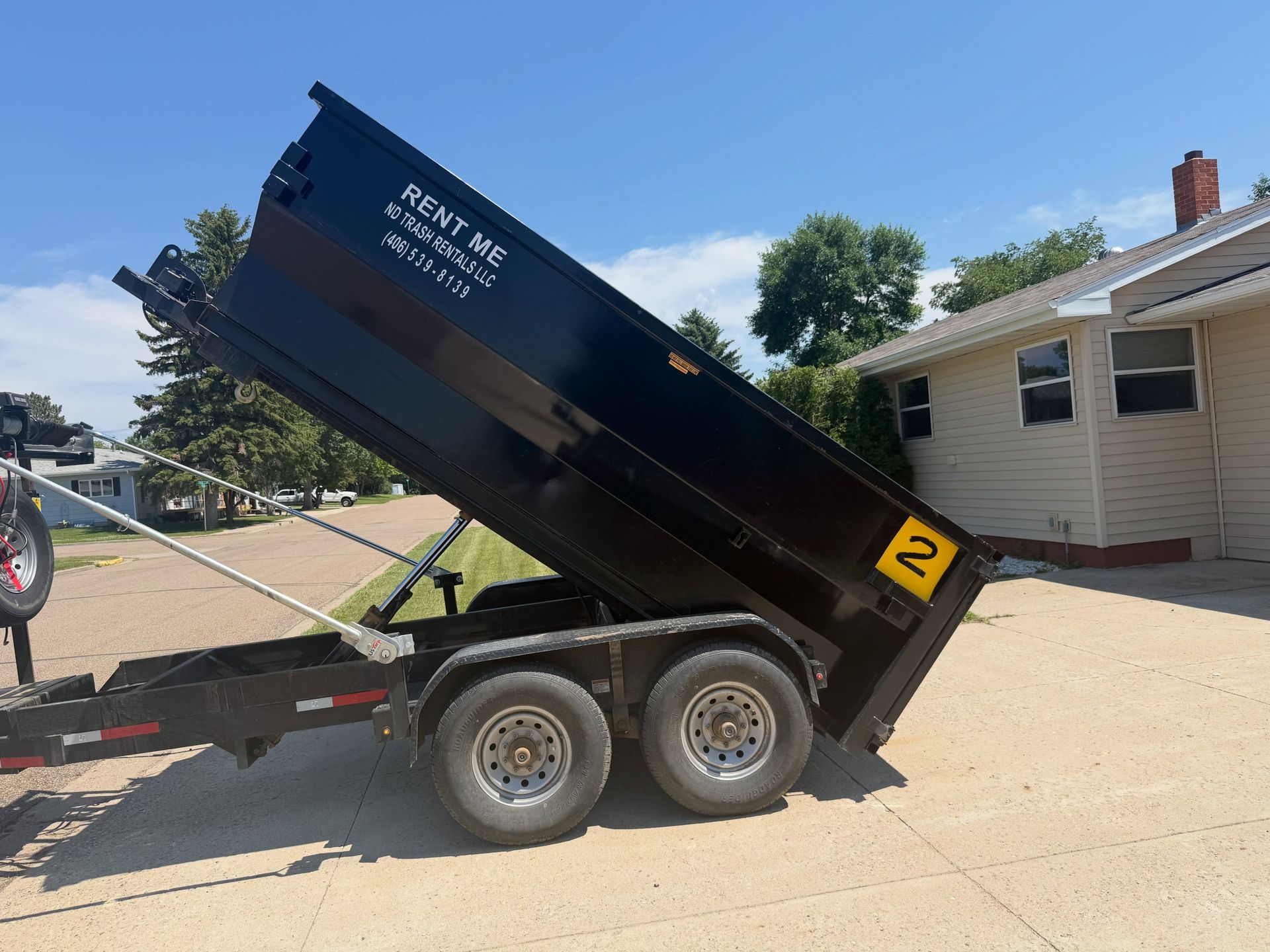 Black dump trailer tipped up on a dual-axle trailer beside a house and driveway under a clear sky