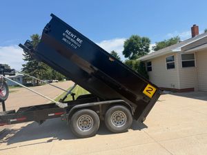 Black dump trailer tipped up on a dual-axle trailer beside a house and driveway under a clear sky