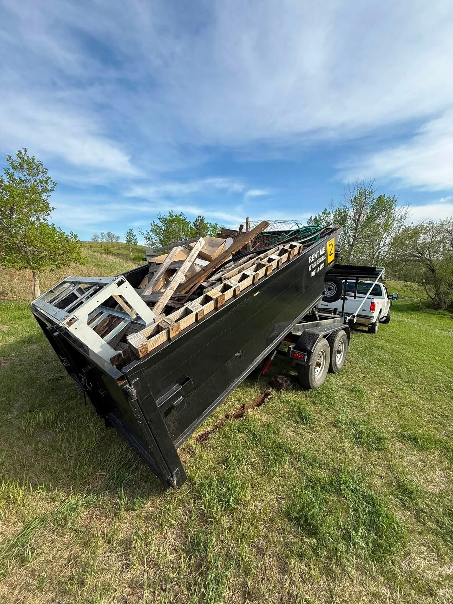 Dump trailer tipped on its side in a grassy field under a blue sky, with a pickup truck nearby.