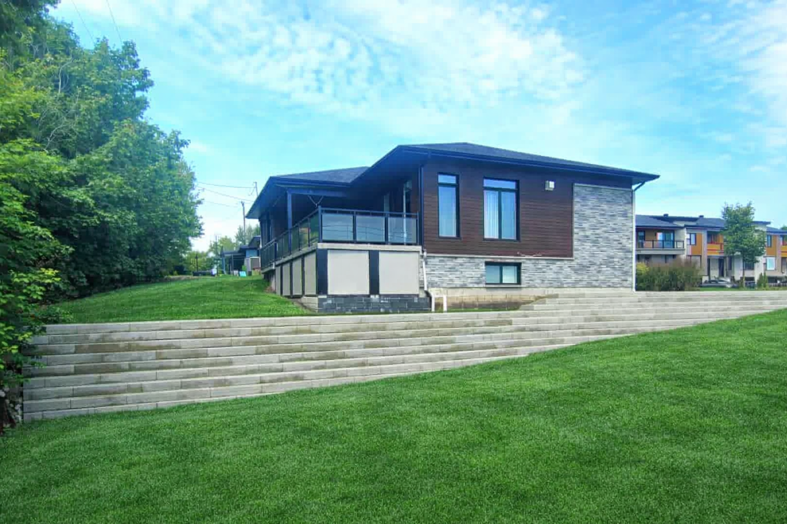 Maison moderne avec extérieur en pierre et en bois, larges marches et cour en pente herbeuse sous un ciel bleu