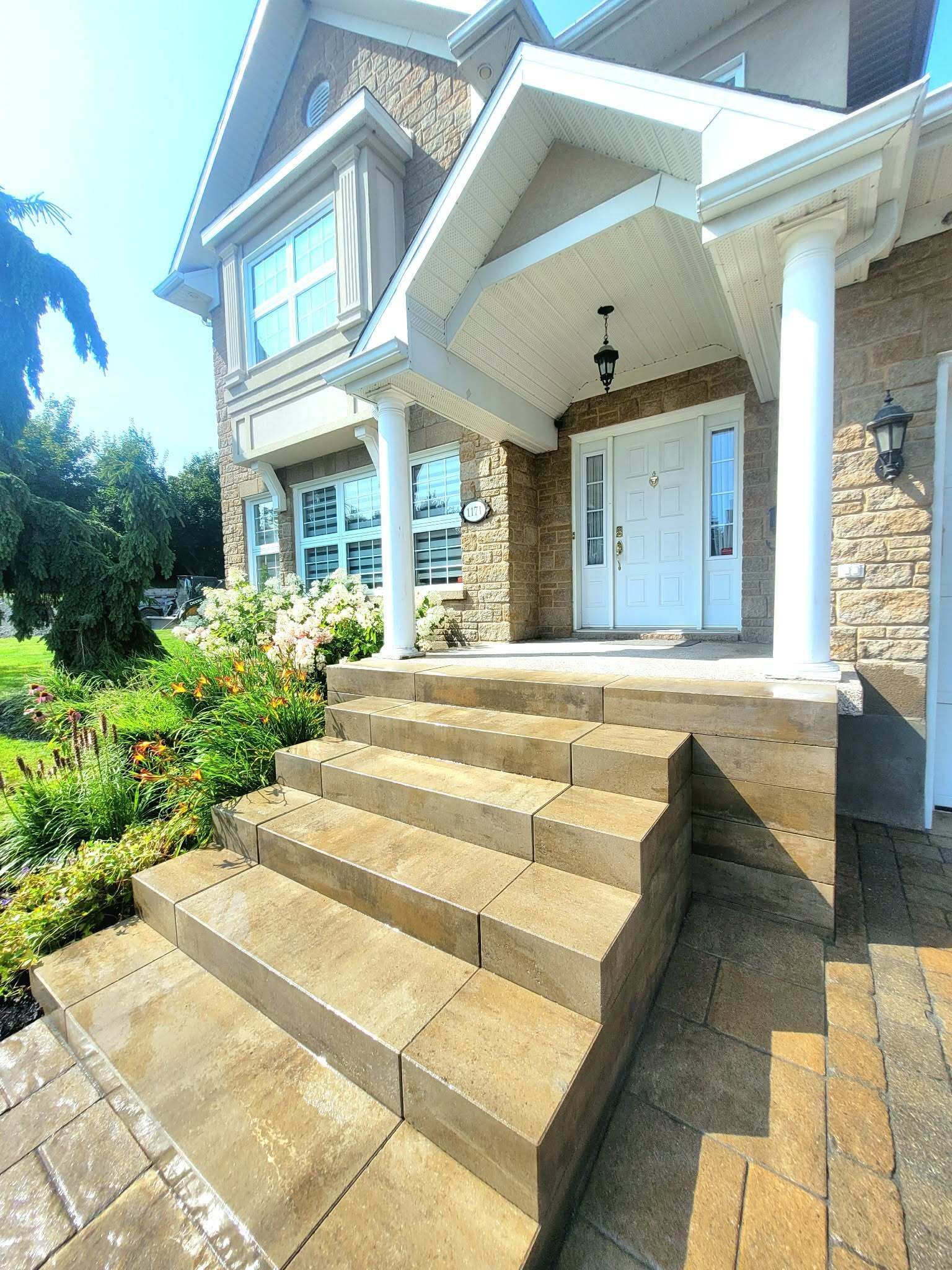 Entrée principale d'une maison avec des marches en pierre beige, des colonnes blanches et un jardin paysager.