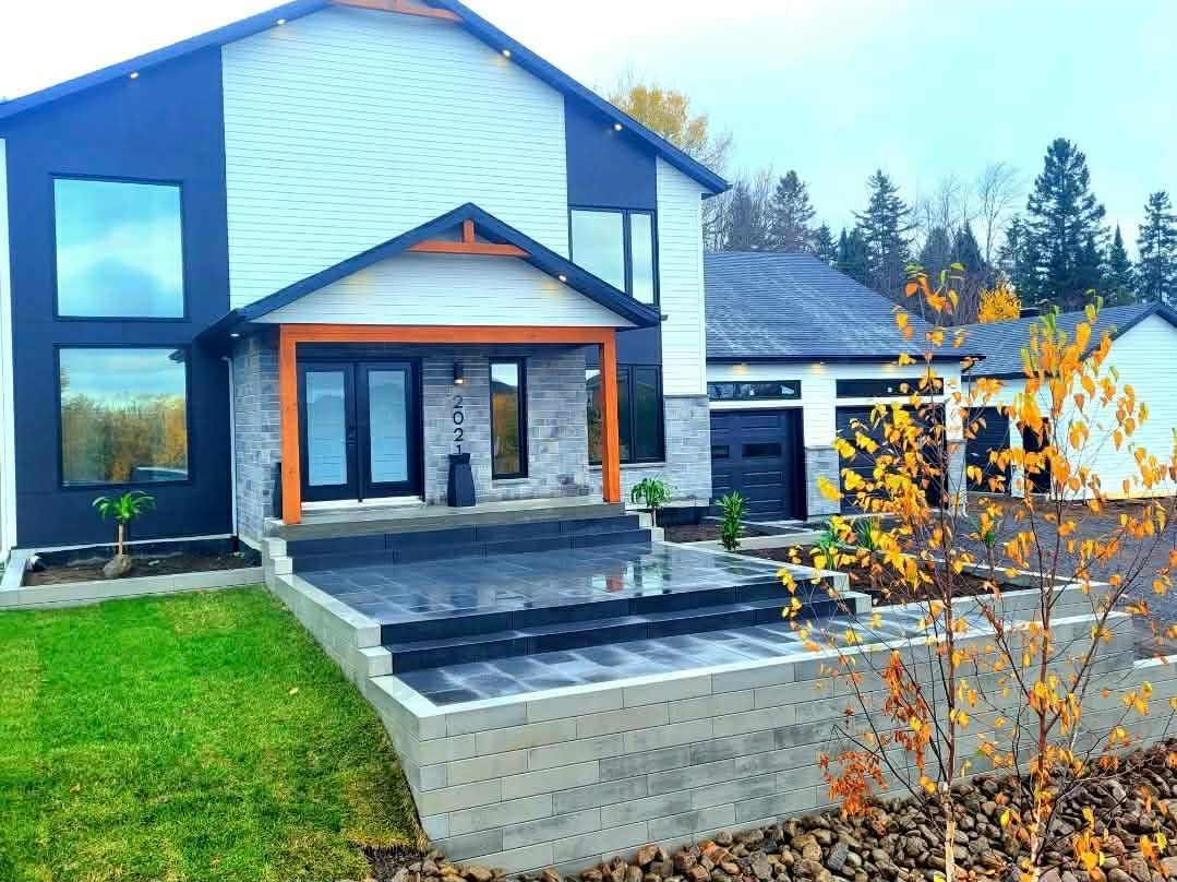 Maison moderne avec façade en bardage foncé, éléments en bois et escalier menant à une terrasse dans un jardin paysager.
