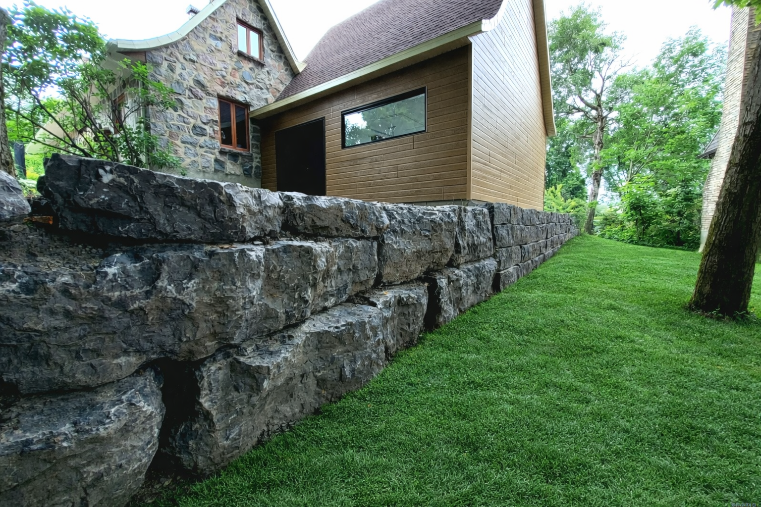 Mur de soutènement en pierre jouxtant une maison brune sur un versant herbeux arboré.