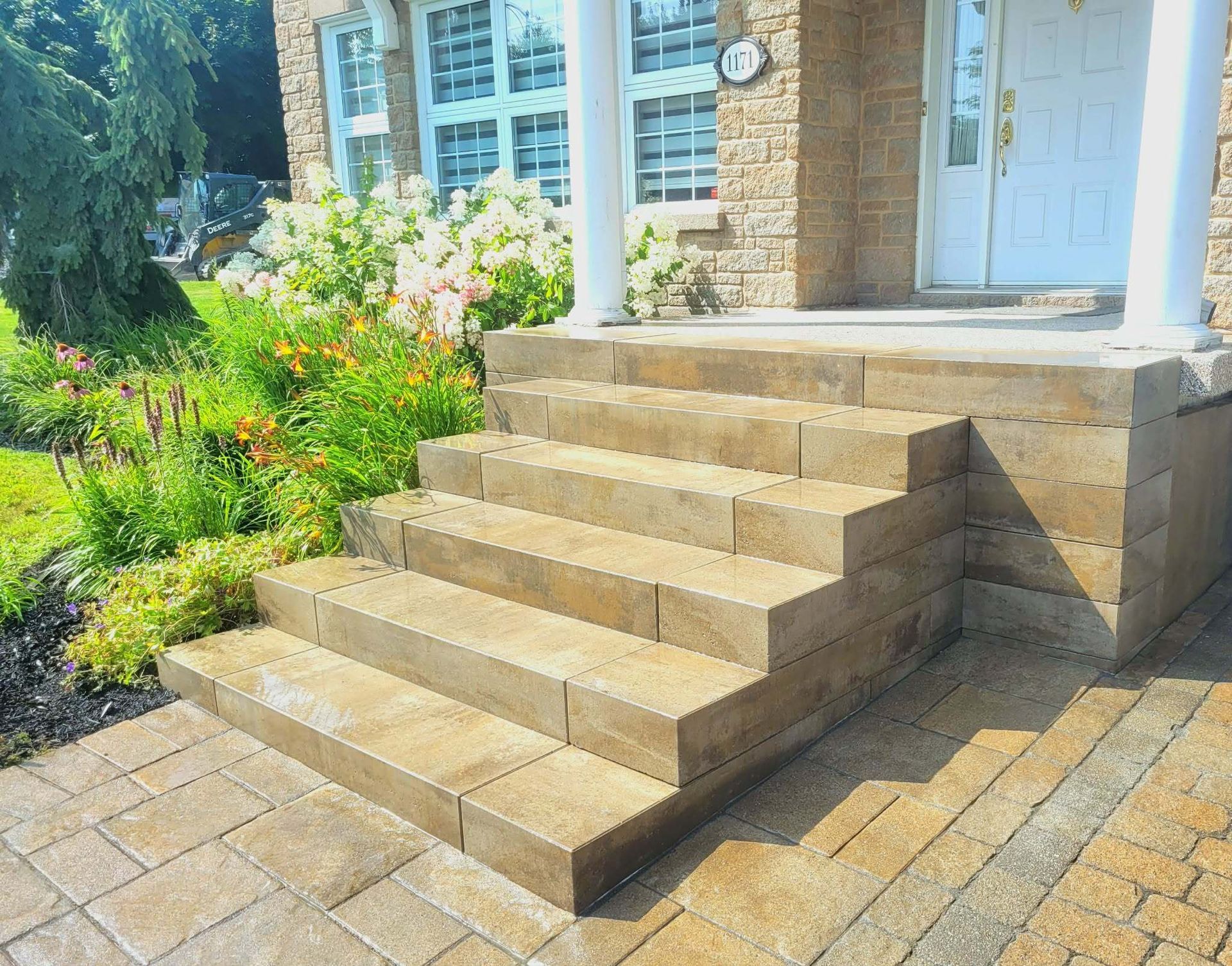 Un escalier en briques mène à une porte blanche donnant sur un jardin ensoleillé et une allée pavée.