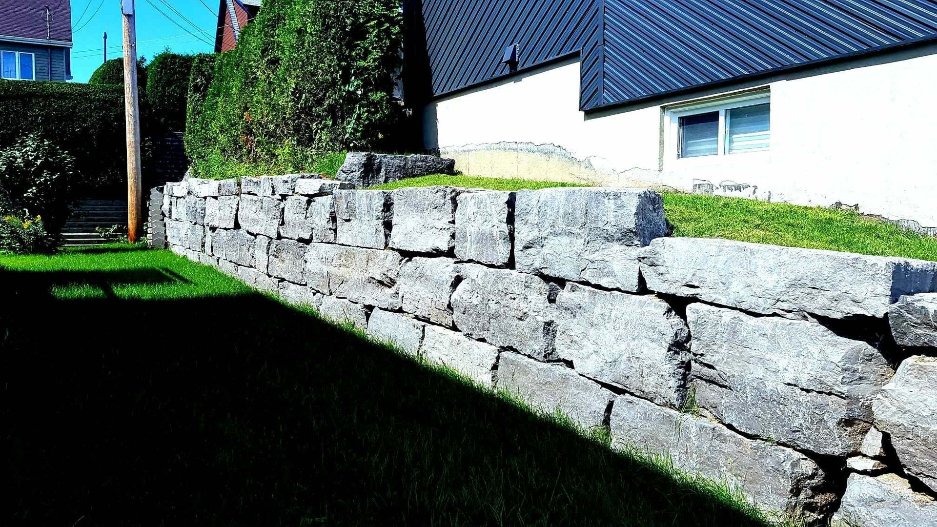 Mur de soutènement en pierre le long d'une cour gazonnée à côté d'une maison baignée de soleil.