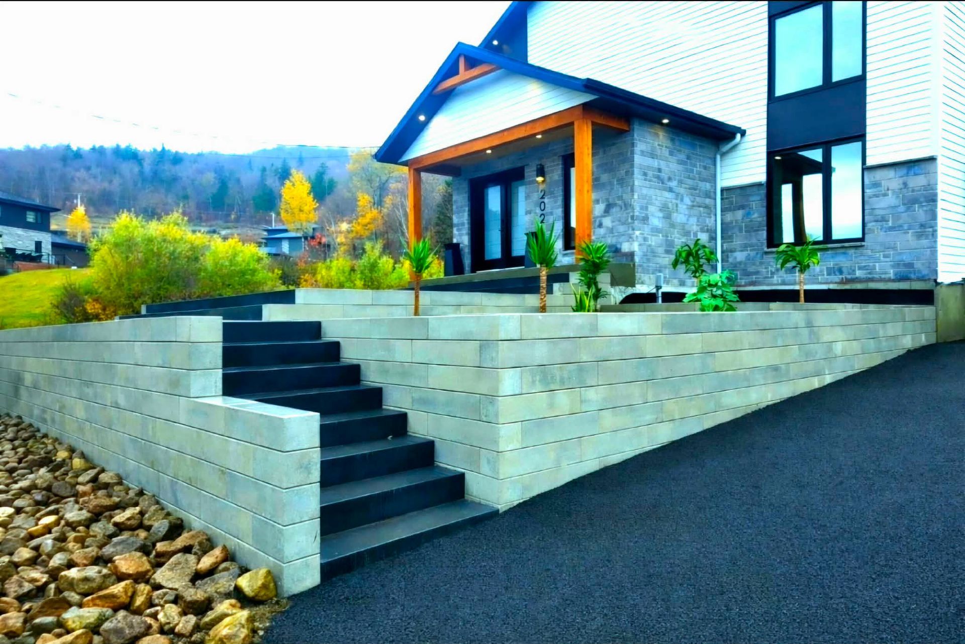 Entrée de maison moderne avec mur de soutènement en pierre, marches et allée en pente dans un jardin paysager.