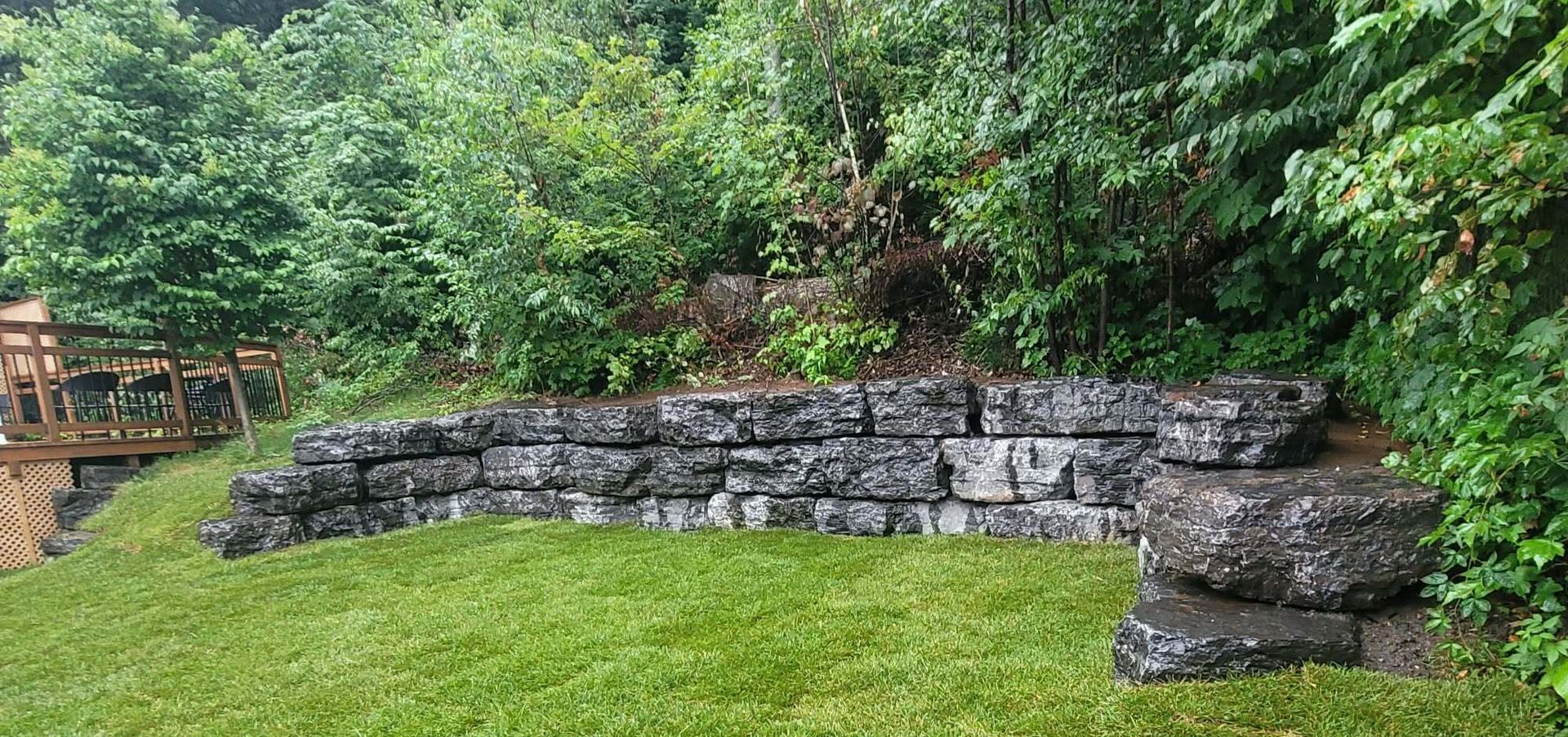 Mur de jardin incurvé en pierres empilées jouxtant une pelouse verte et des arbres denses après la pluie