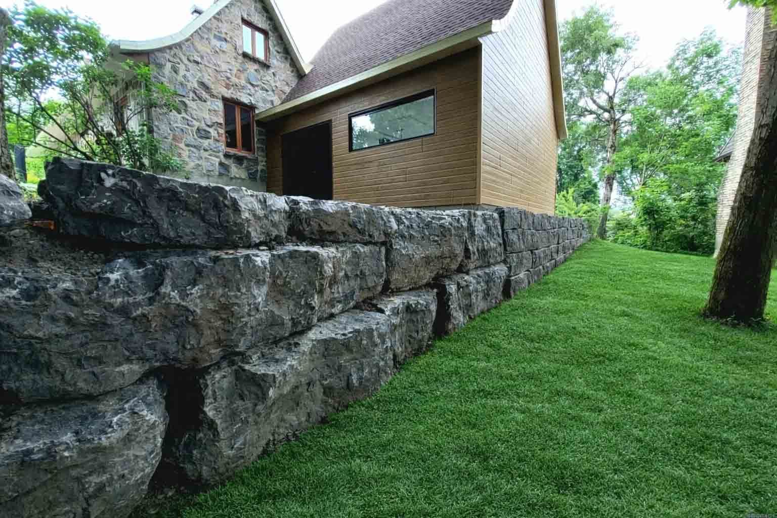 Mur de soutènement en pierre bordant une pelouse verte, avec une maison brune en arrière-plan.