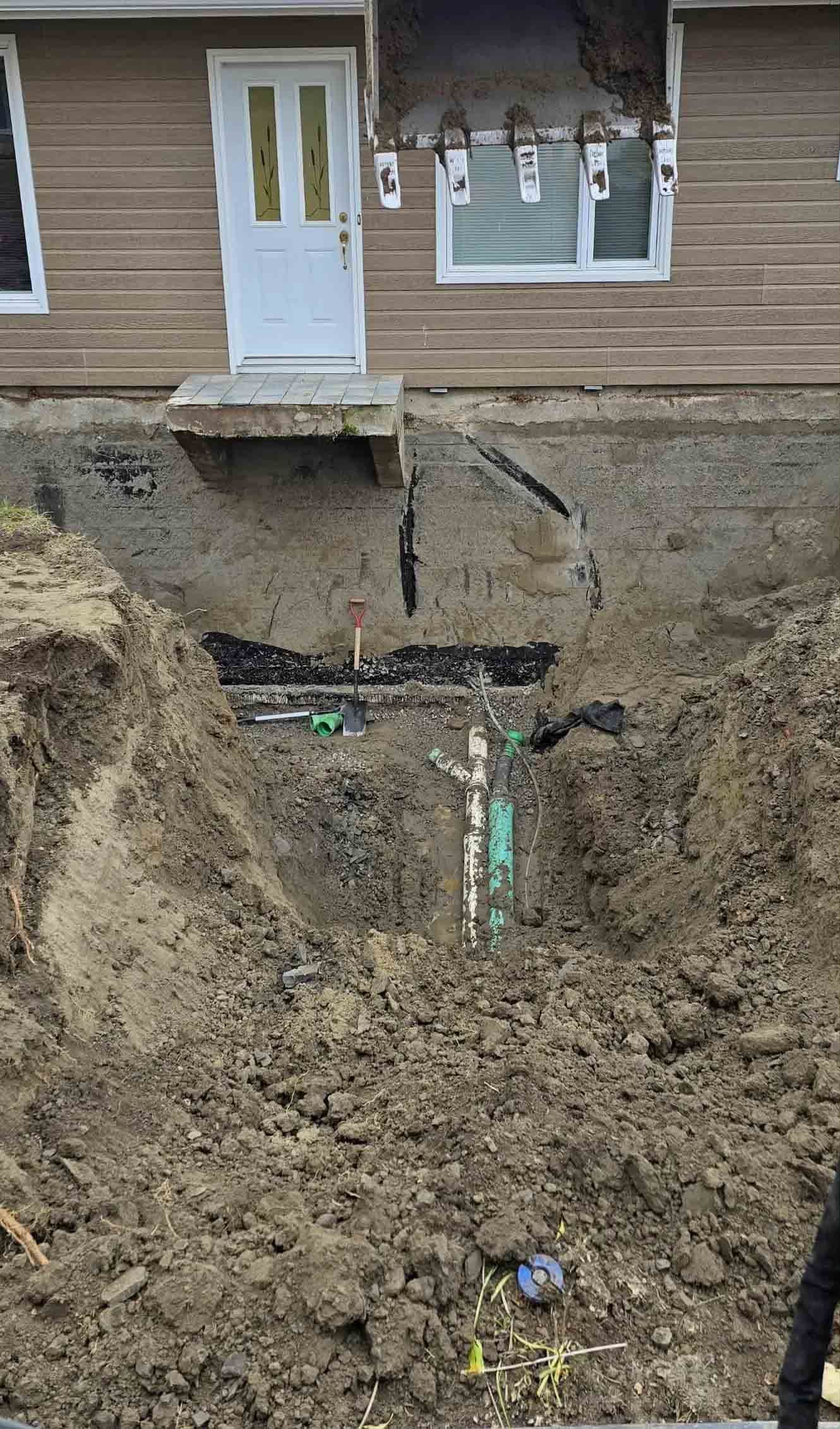 Un ouvrier portant un casque de chantier jaune installe des tuyaux près de fondations recouvertes d'un revêtement noir étanche.