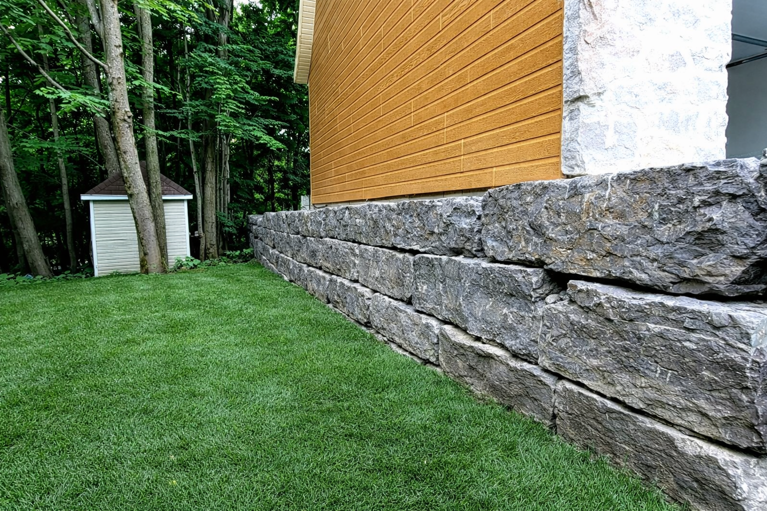 Maison avec fondations en pierre, bardage orange et pelouse verte en bordure d'un bois.