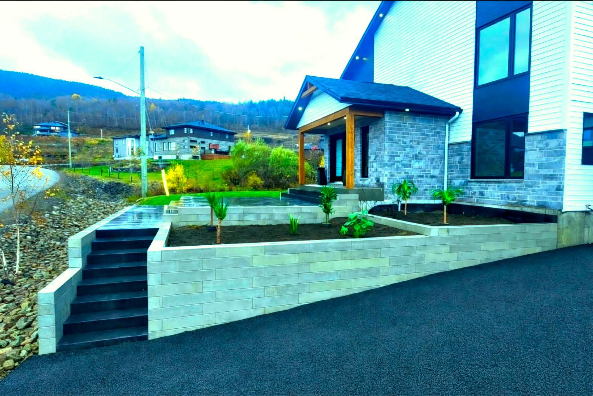 Maison moderne avec escalier en pierre, mur de soutènement et jardin paysager en bordure de route, avec les montagnes en arrière-plan.