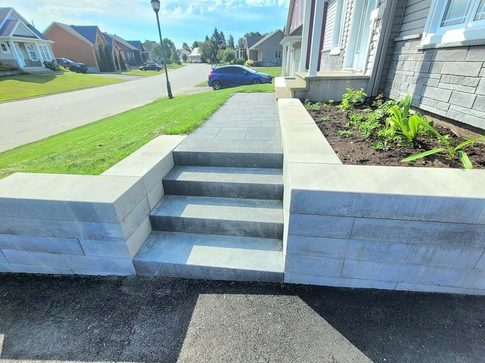 Un escalier et une rampe mènent à l'entrée de la maison, avec des parterres aménagés et une vue sur une rue de banlieue.