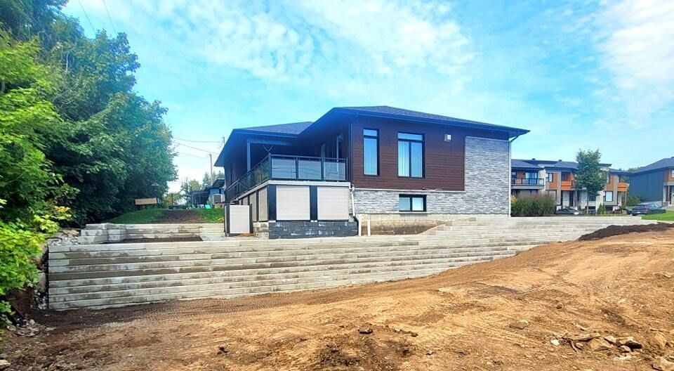 Maison moderne avec façade en pierre et toit en pente sur un terrain en terre battue, vue de la rue sous un ciel bleu