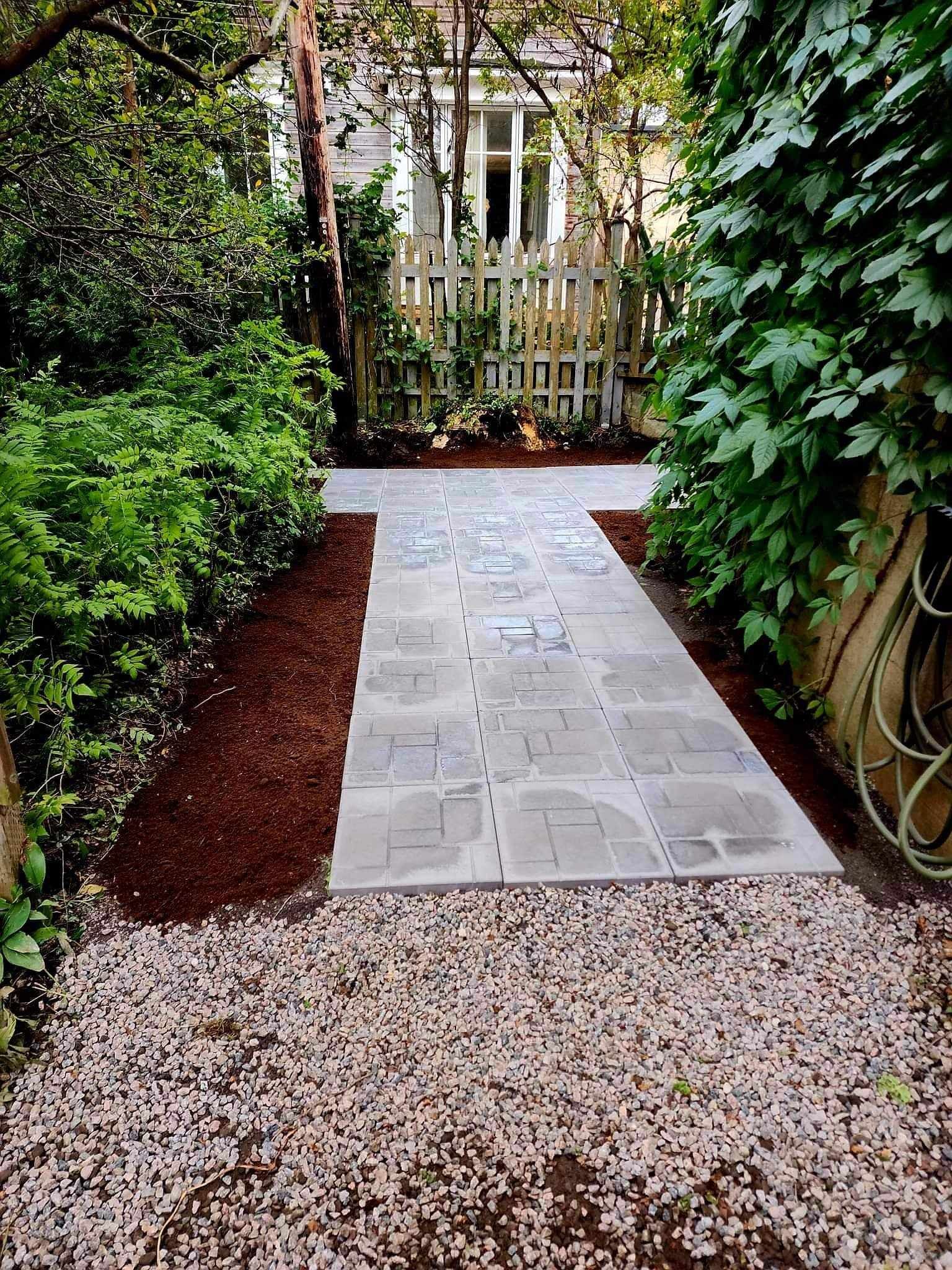 Allée de jardin en pierre bordée d'une végétation dense menant à un portail blanc.