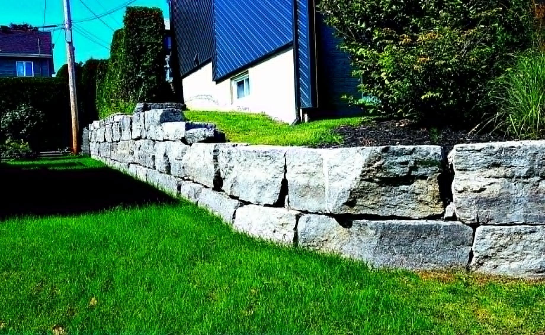 Mur de soutènement en pierre incurvé jouxtant une pelouse verdoyante et une maison baignée de soleil.