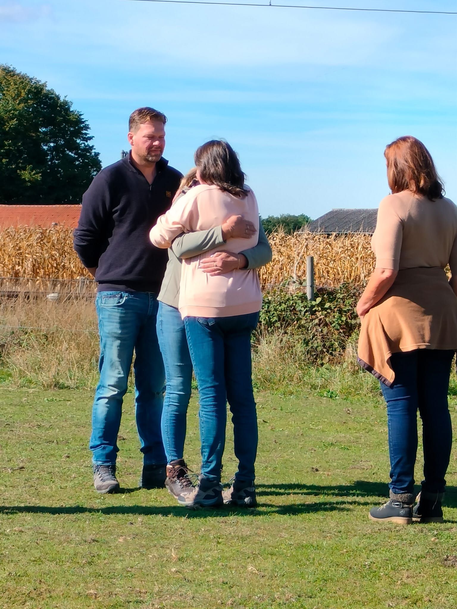 Vier mensen buiten. Twee omhelzen elkaar, de anderen kijken toe. Veld met een blauwe lucht op de achtergrond.