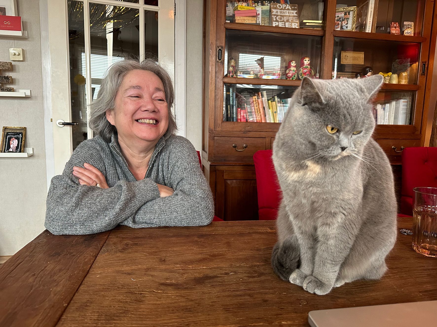 Astrid glimlacht naar een grijze kat; beiden zitten aan een houten tafel voor een boekenkast.