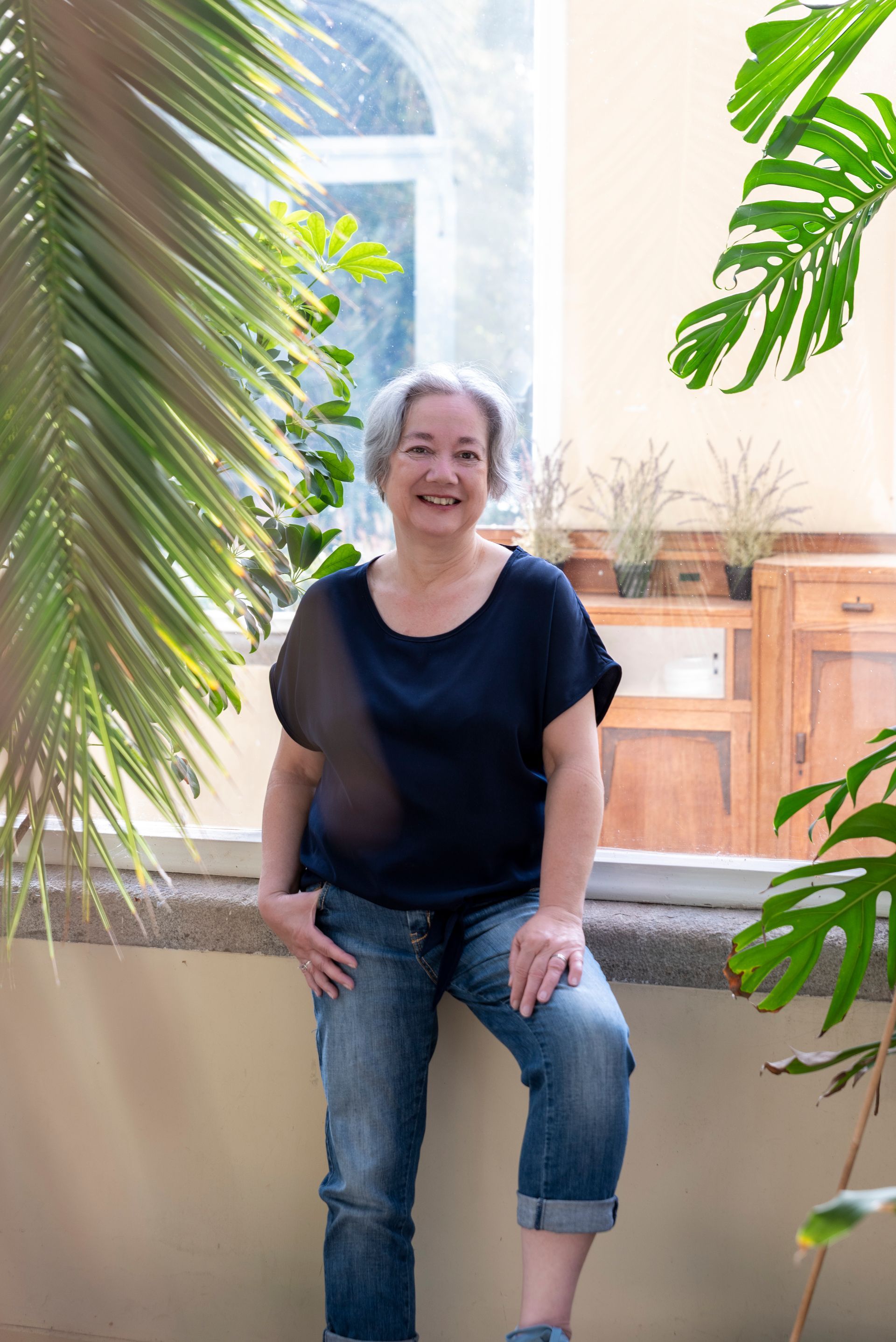 Een vrouw in een blauwe blouse en spijkerbroek staat lachend tegen een raam in een zonnige kamer vol planten.