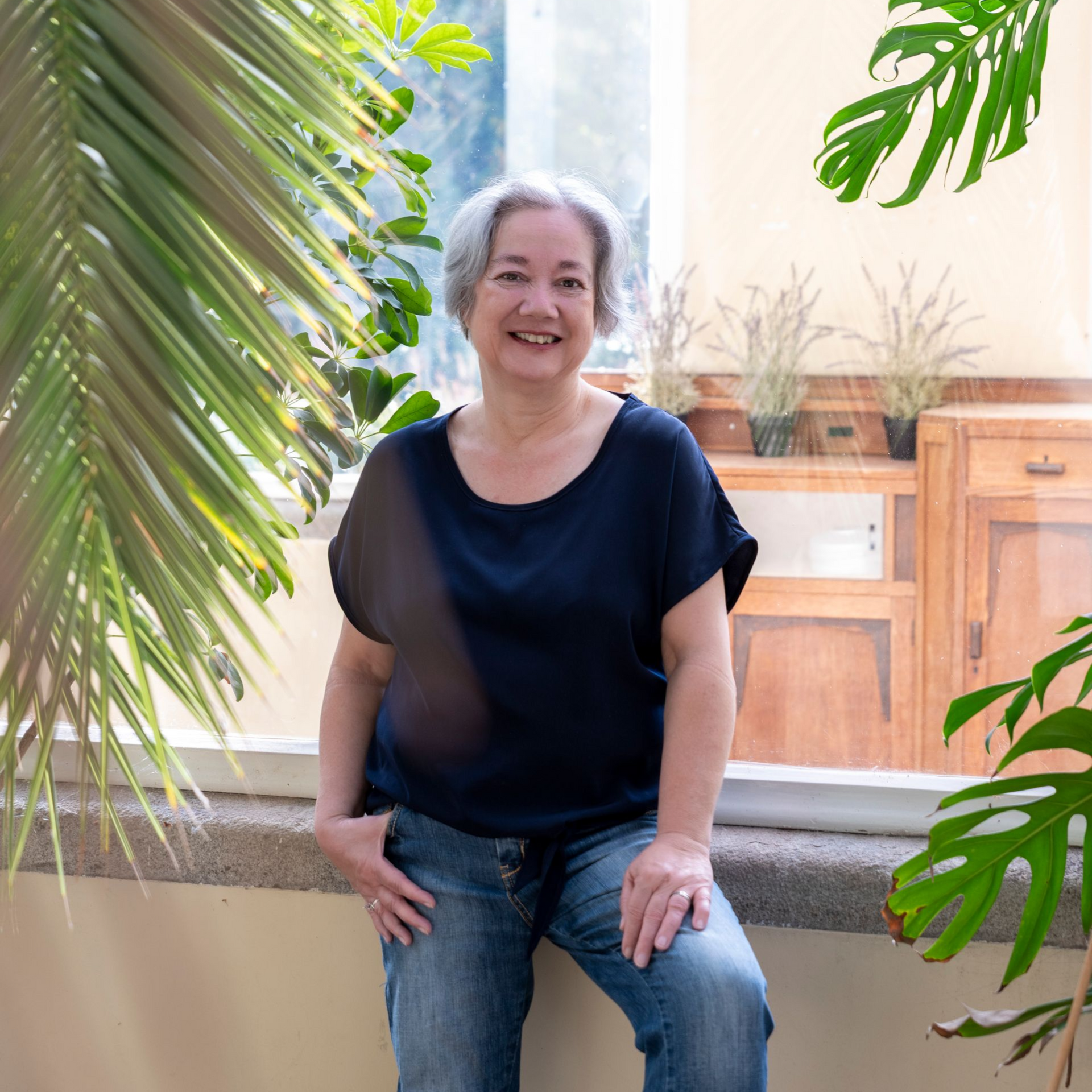 Astrid staat voor een raam tussen de planten. Ze draagt een blauwe blouse, rechthand in de zak, linkerhand op haar knie.