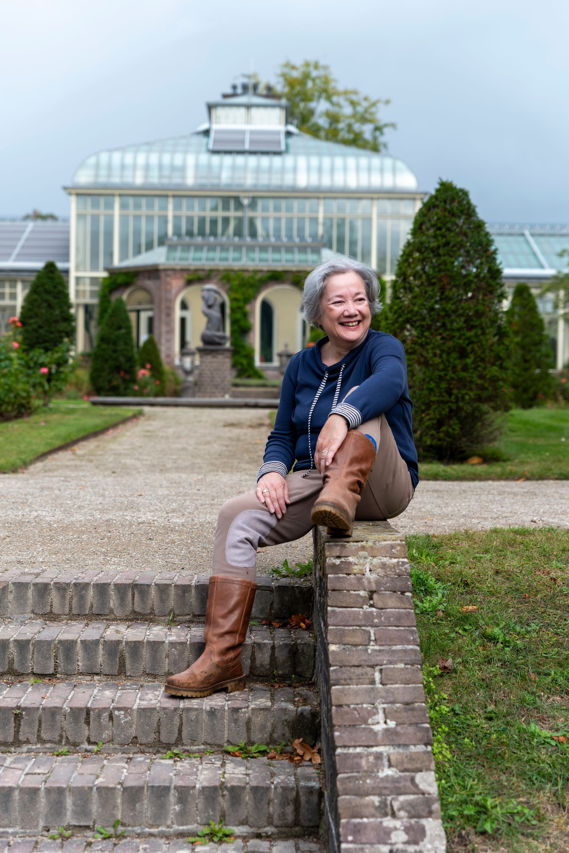 Een vrouw in laarzen zit glimlachend op een bakstenen muur voor een glazen gebouw en tuin.