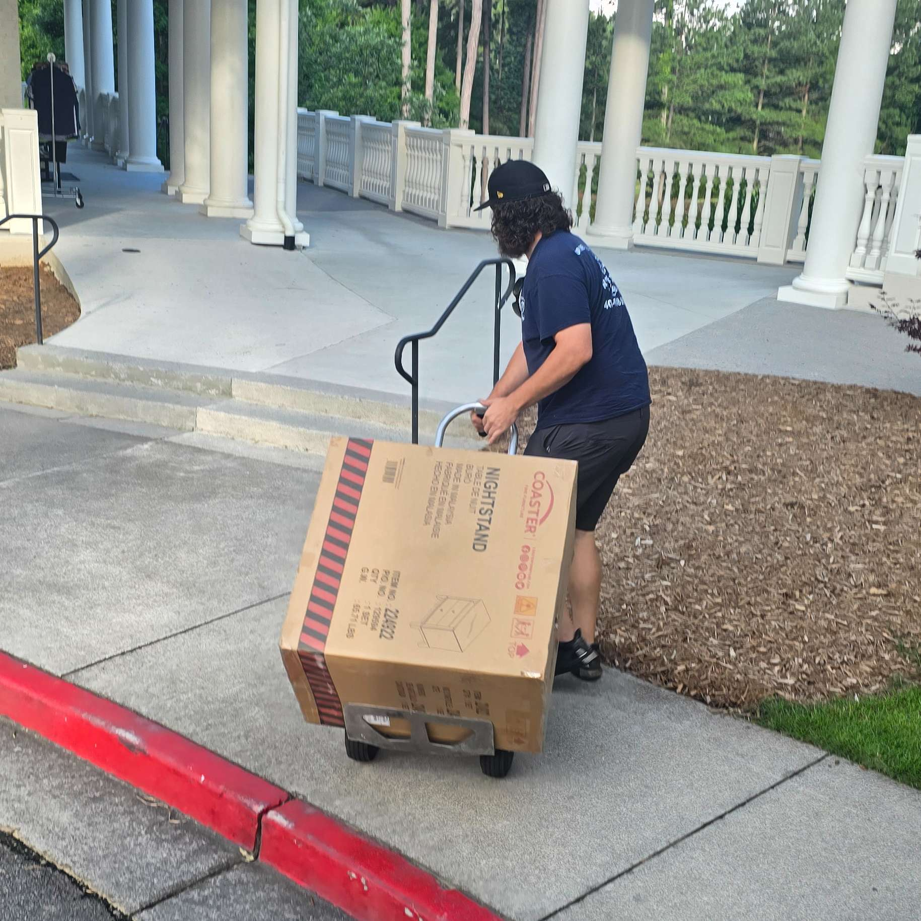Person pulling a large cardboard box on a hand truck up a slight incline. Building with columns in background.