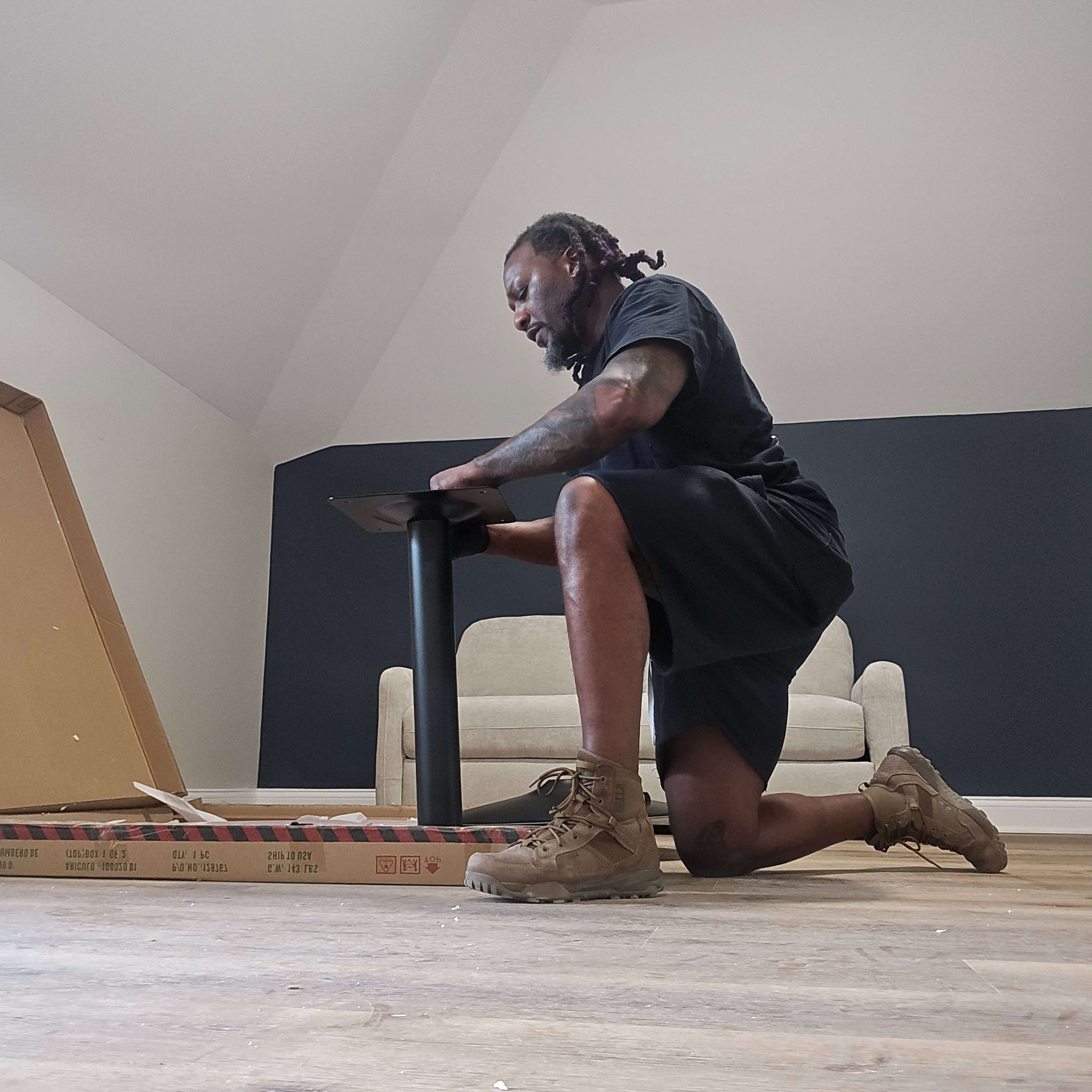 A person assembling furniture on their knees, indoors. They're wearing dark clothing and boots. Boxes are nearby.