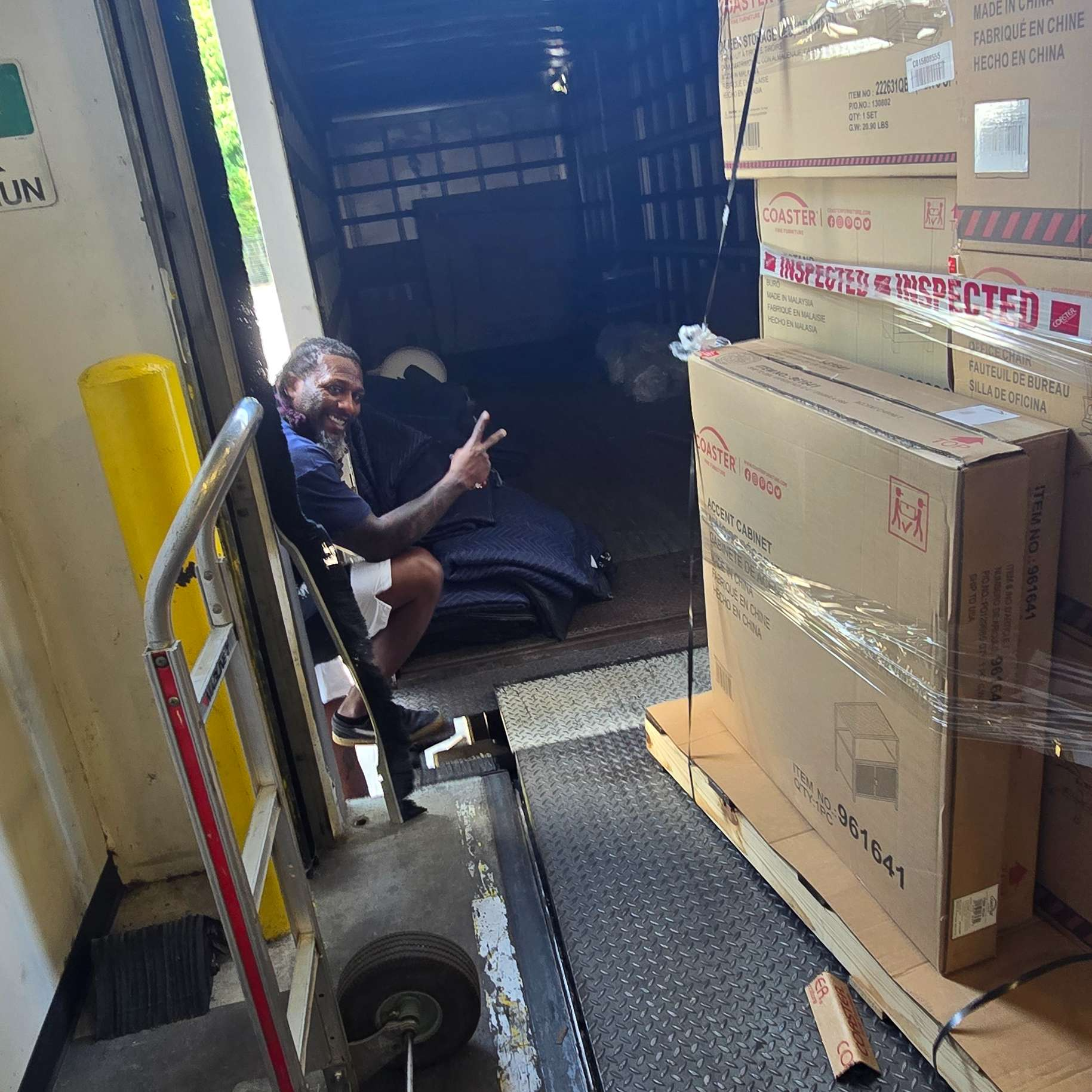Man sits in a truck, surrounded by boxes and a dolly, giving a peace sign.