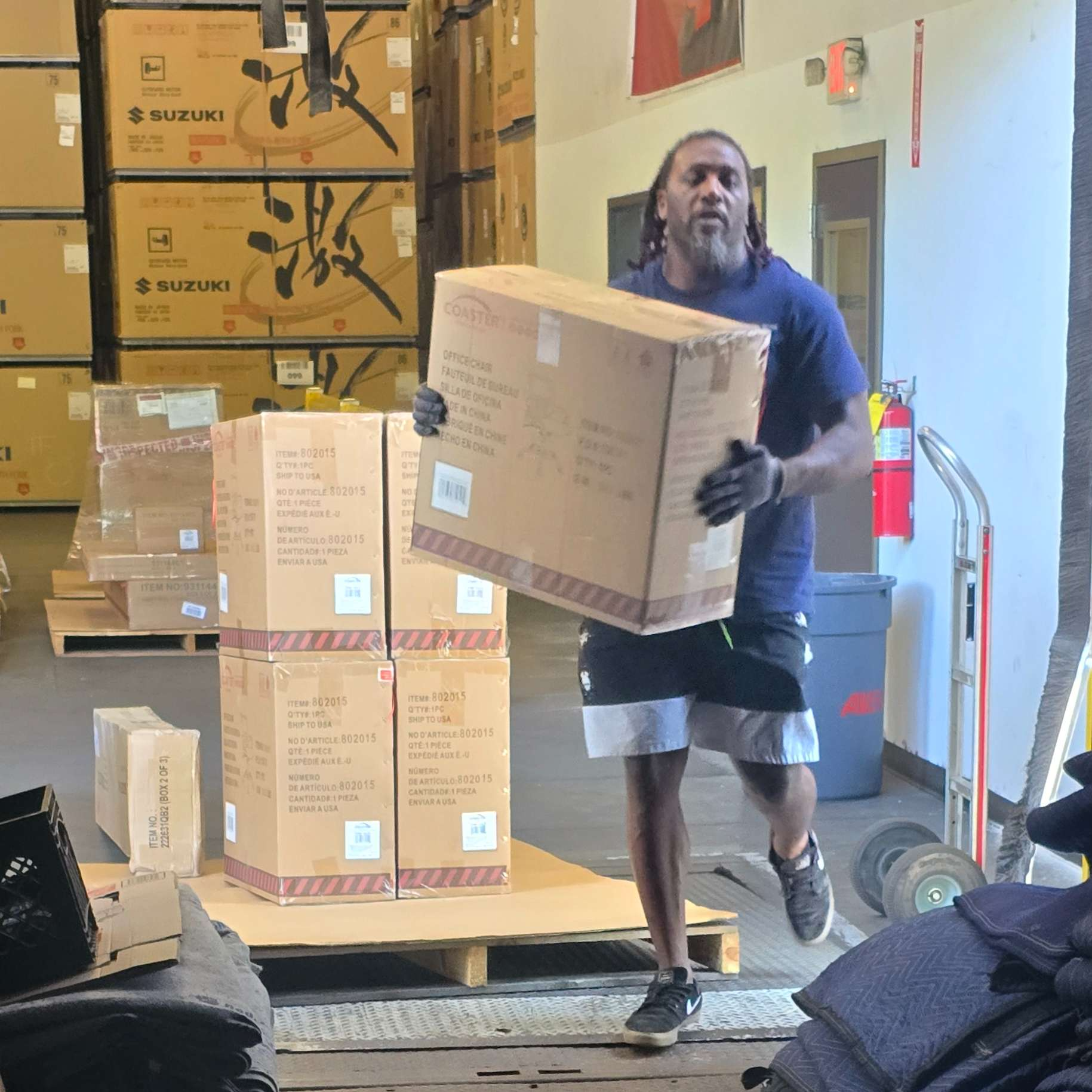 Man carrying a box in a warehouse, with boxes stacked on a pallet nearby.