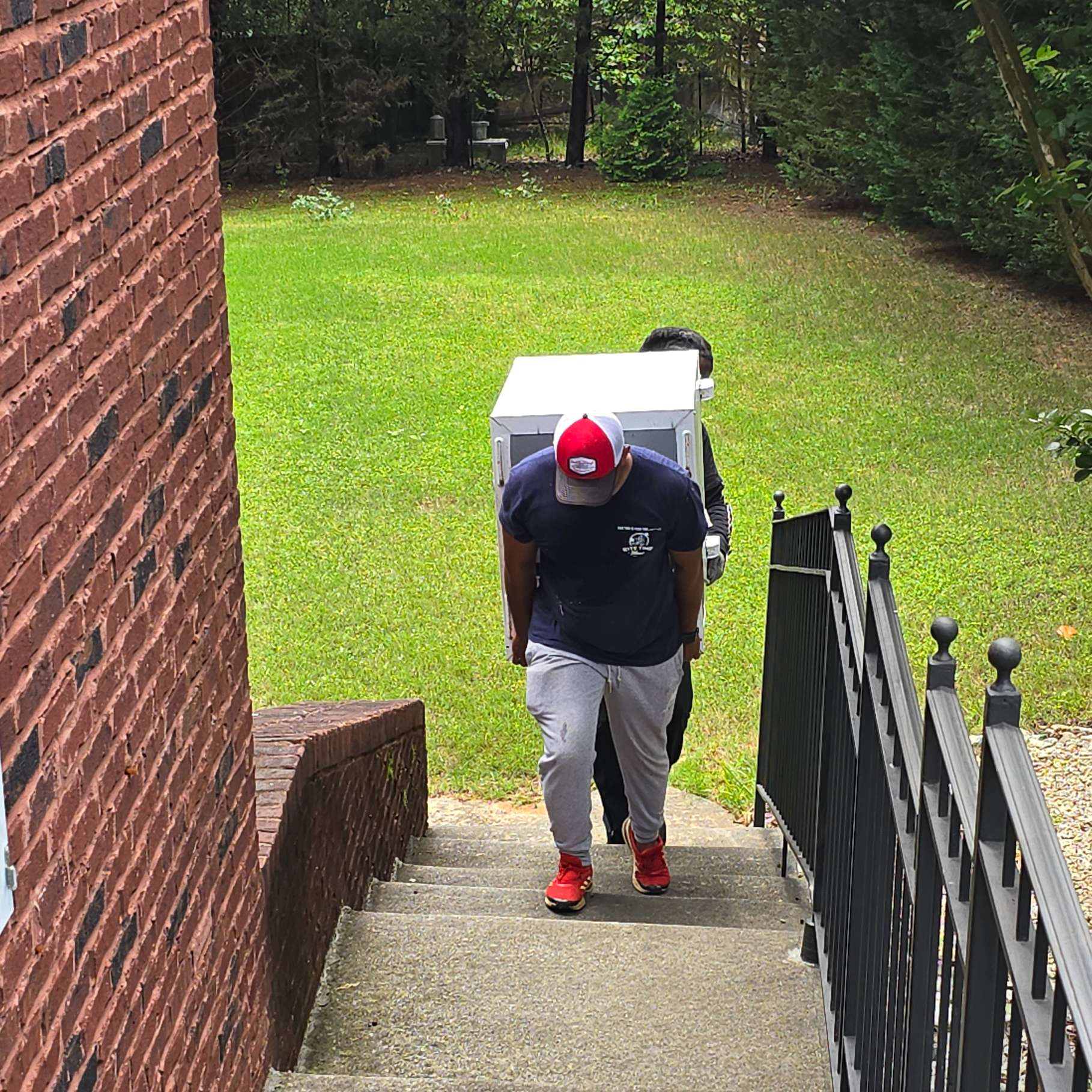 Two people carry a white box up outdoor concrete stairs. A red brick wall is on the left.