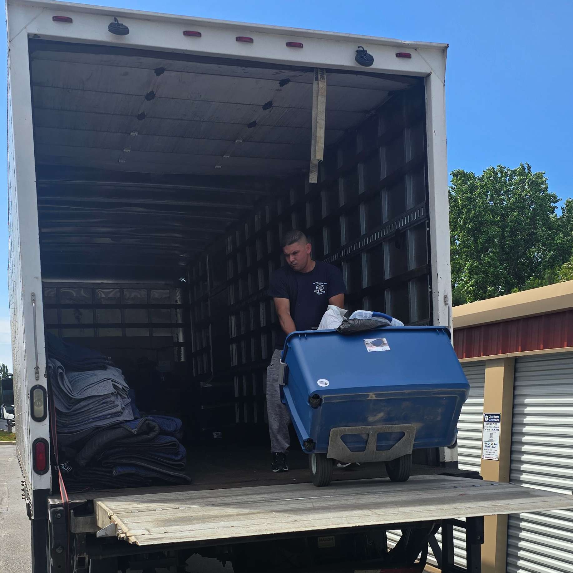 Man moving a blue bin on a dolly into the back of a moving truck. The truck is open, revealing the empty interior.