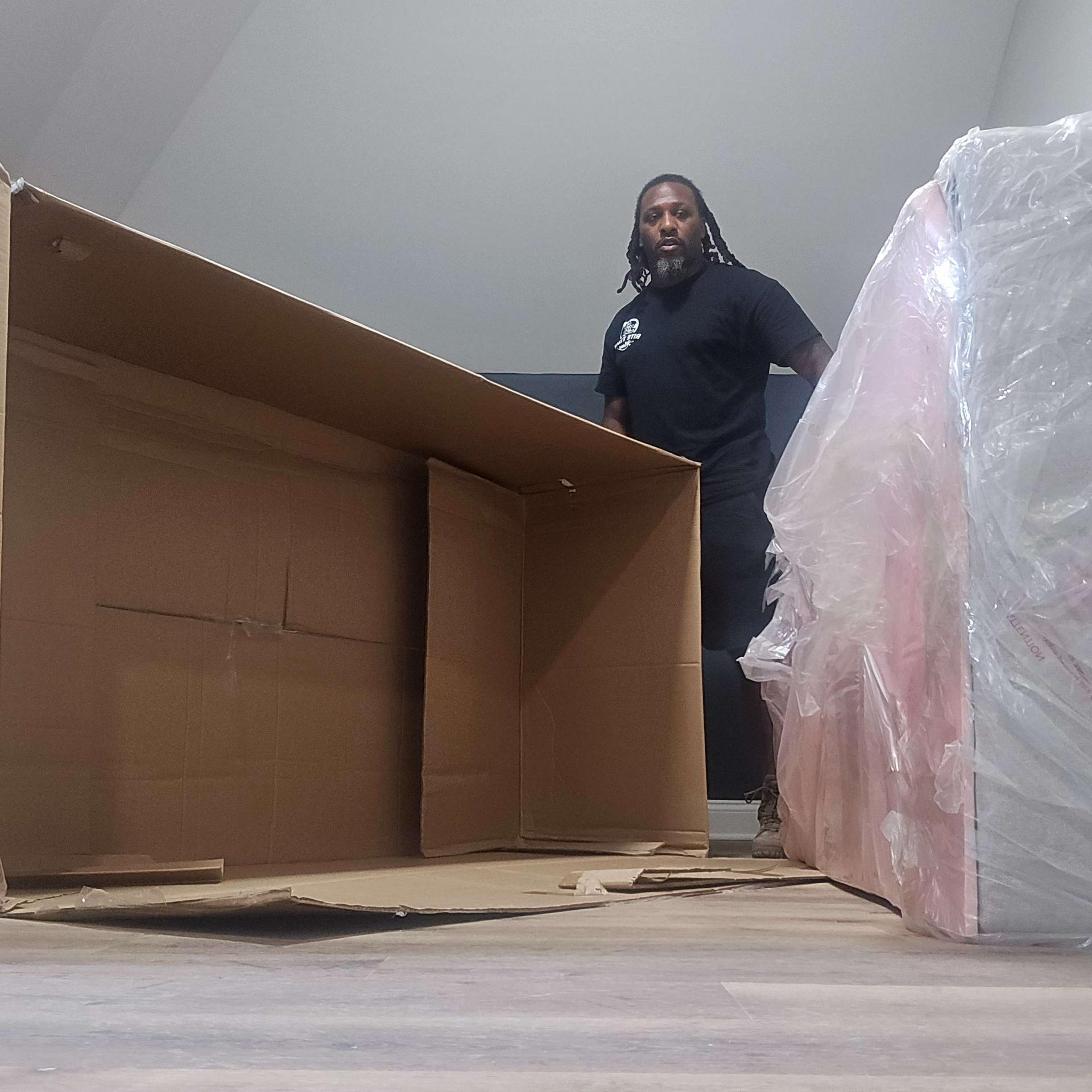 A person in black clothing standing near a large, open cardboard box and a wrapped object, indoors.