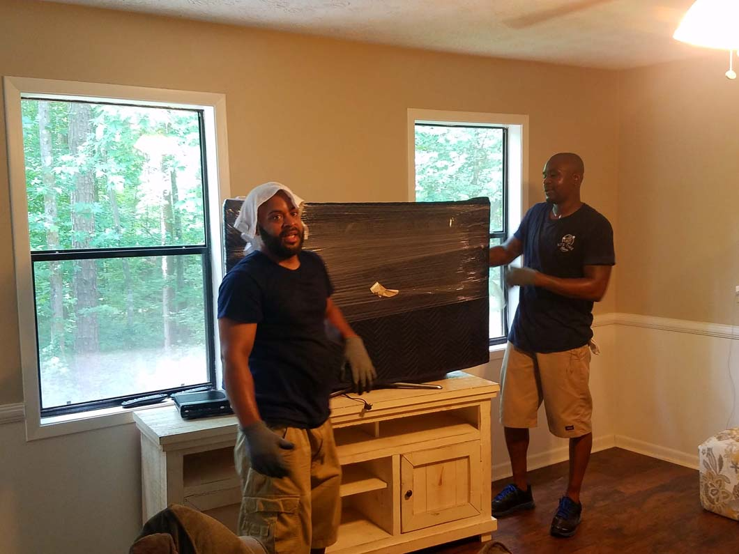 Two men moving a wrapped TV. One in a head covering smiles. Wooden cabinet, windows.