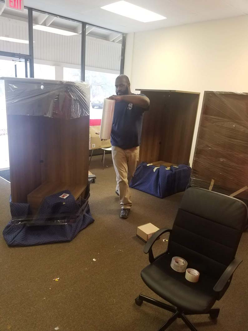 Man carrying box in office; furniture wrapped in protective coverings.