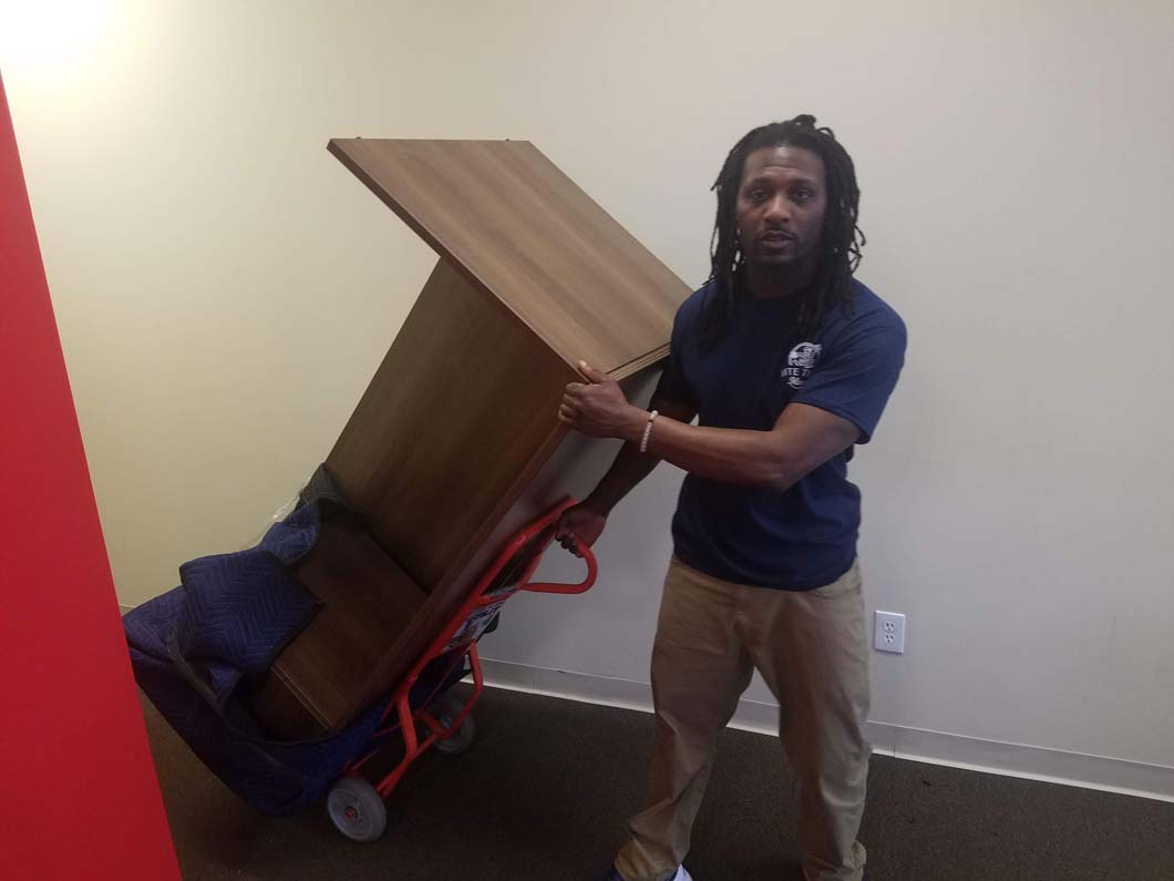 Man moving a brown table with a hand truck against a beige wall, red accent.