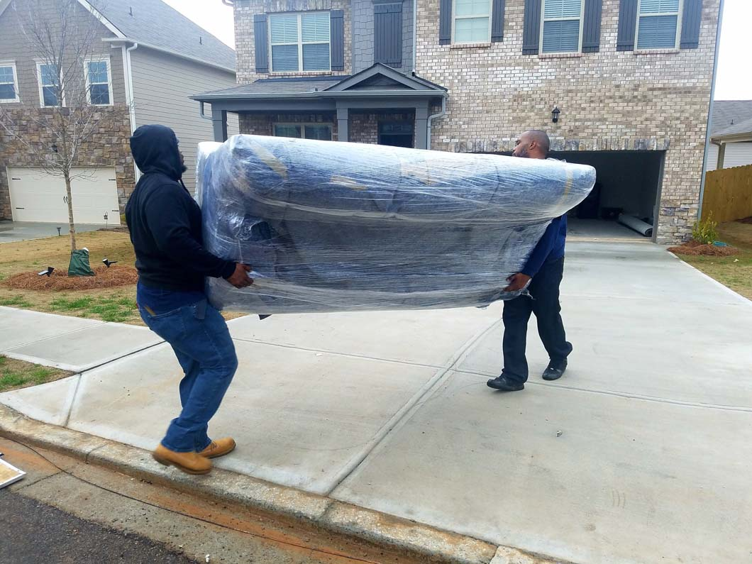 Two people carrying a large, wrapped sofa on a driveway in front of a house.
