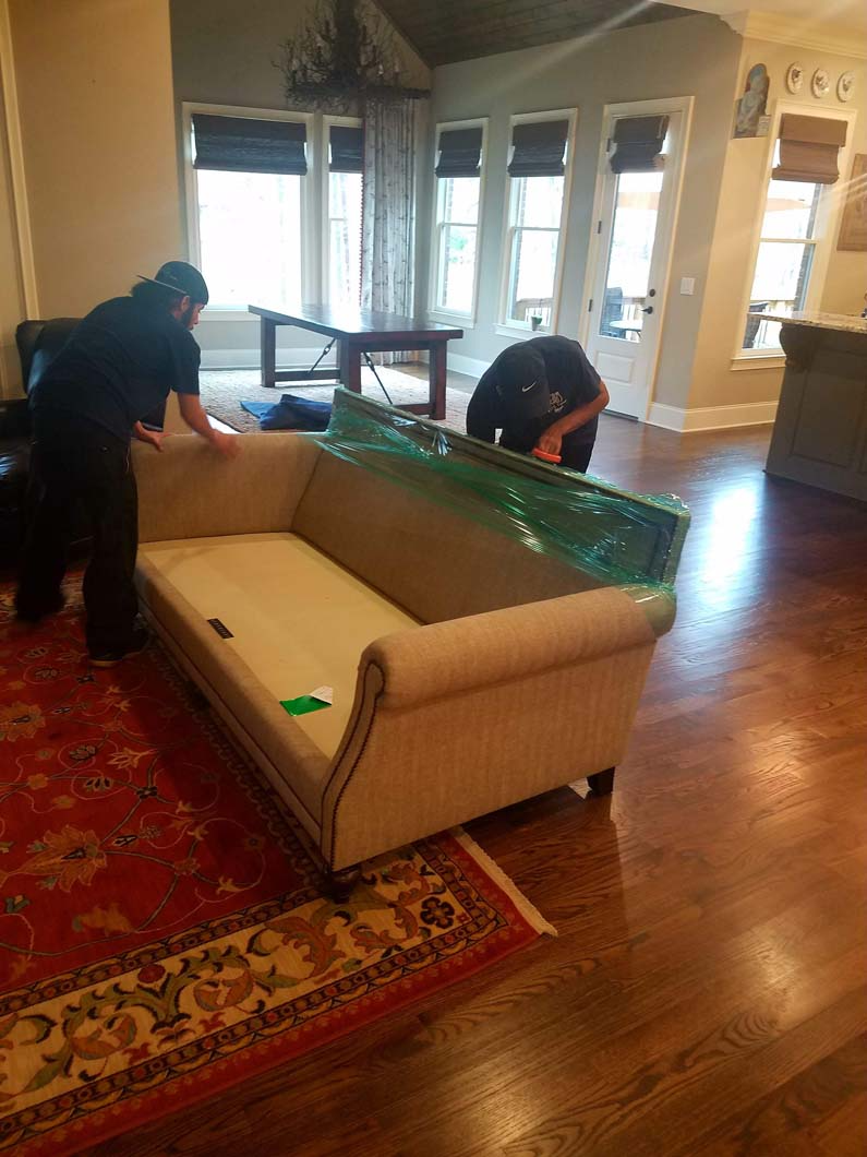 Two people wrapping a beige sofa in plastic in a living room with wood floors and a red rug.