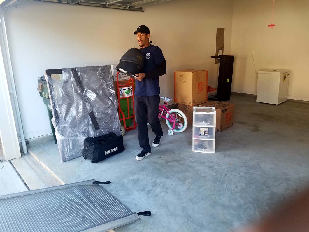 Man in a garage holding a helmet, surrounded by packed boxes, furniture, and a bicycle.