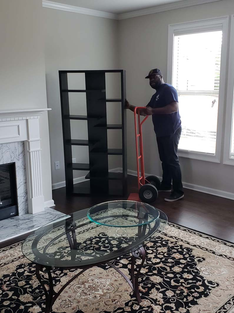 Man moving a tall black shelf with a dolly in a living room; fireplace and glass coffee table in view.