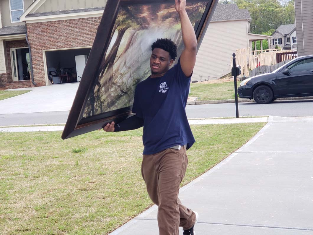 Person carrying a large framed painting outdoors on a sunny day.