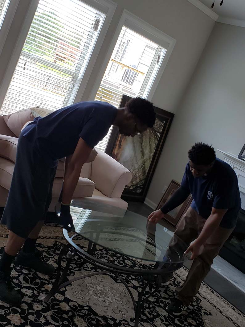 Two people lifting a glass-topped table indoors; a couch and window in the background.