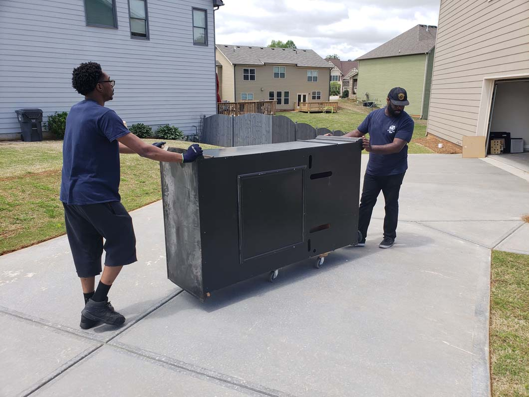 Two people pushing a large, dark cabinet on wheels across a concrete driveway.