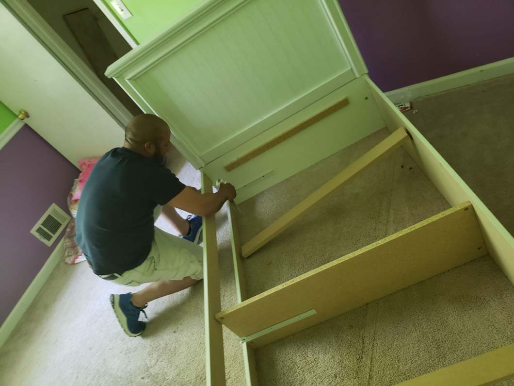 A person assembling a white and beige storage bed on a carpeted floor in a room with purple and green walls.