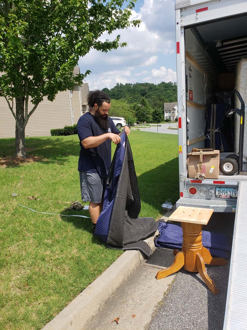 Man packing items into a moving truck. Includes a side table and furniture padding on grass.