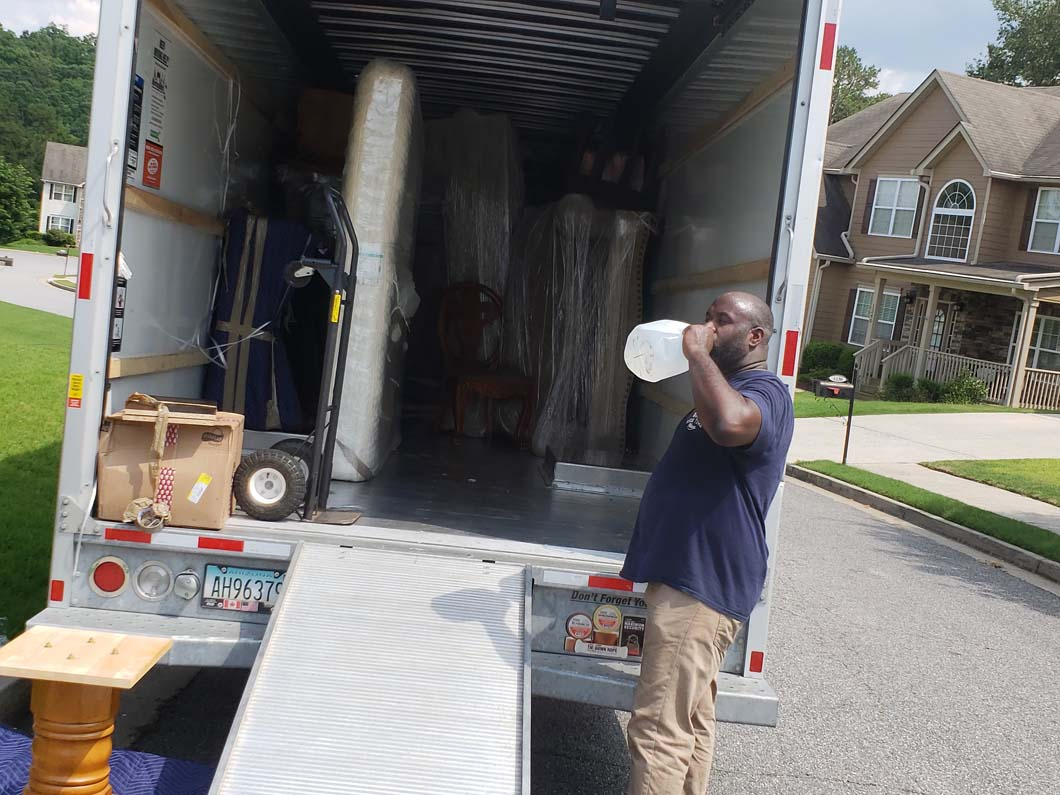 Man drinking water near an open moving truck loaded with furniture on a driveway.