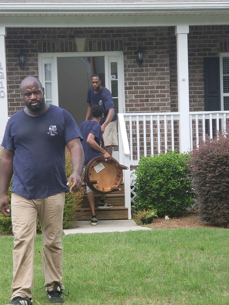 Movers carrying furniture out of a house; one man walks towards the viewer.