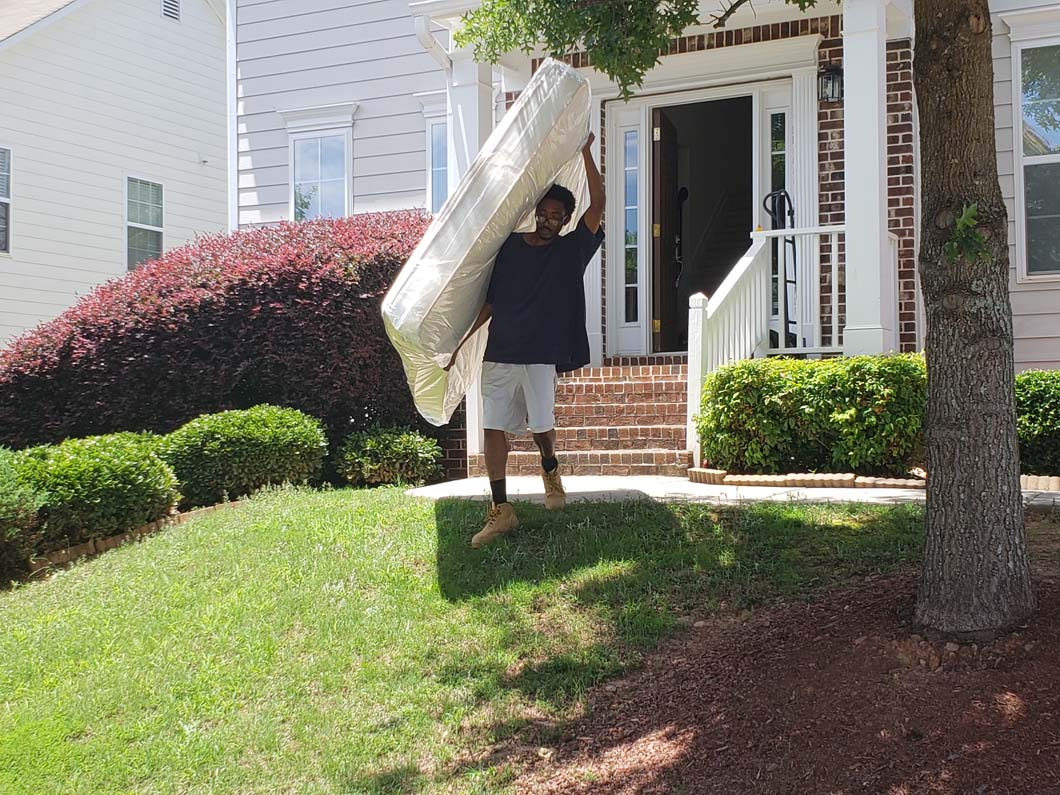 Person carrying a mattress toward a house with a white door and green lawn.