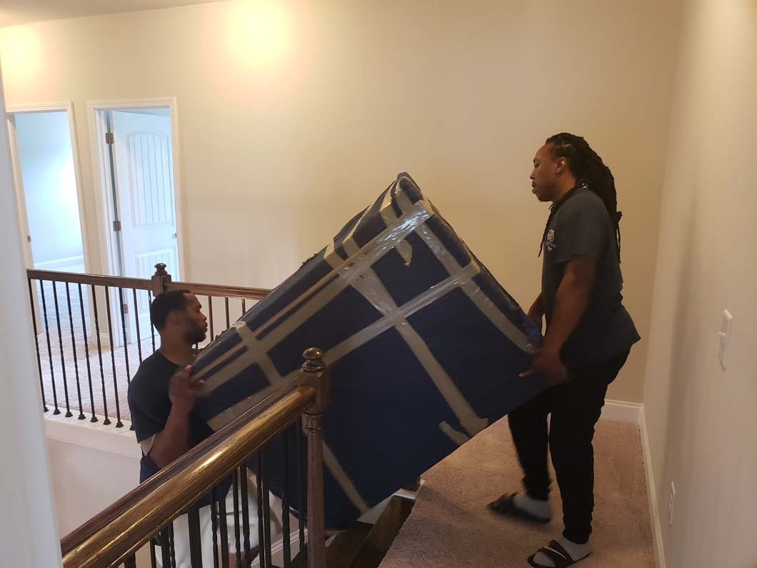 Two people carrying a large, wrapped item up carpeted stairs with a railing.